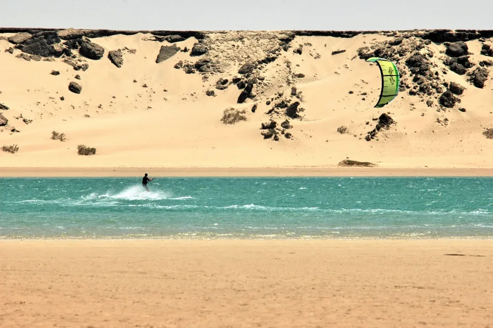 Un kitesurfeur file sur l’eau turquoise de la lagune de Dakhla, au pied d’une dune de sable clair.